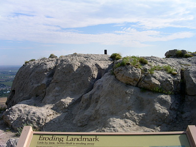 [At the bottom of the image is the top edge of the sign labeled Eroding Landmark. Across the way is the top of the bluff with a cylindrical metal pipe sticking out from the top of the rock by probably at least a foot.]